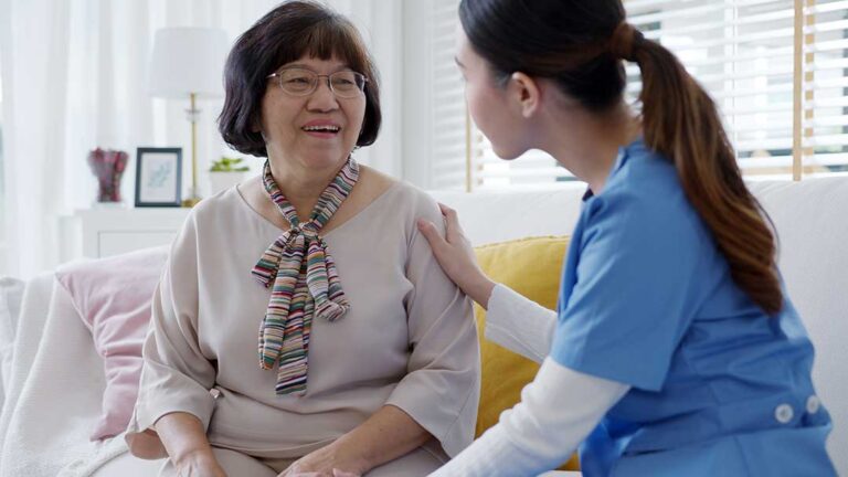 An older adult receives daily medication from a caregiver with organized pill containers on a kitchen table.